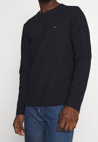 Schwarzes Langarm-T-Shirt aus strukturiertem Stoff, Rundhalsausschnitt und kleinem rot-weiß-blauem Logo auf der Brust. Kombiniert mit blauen Jeans.