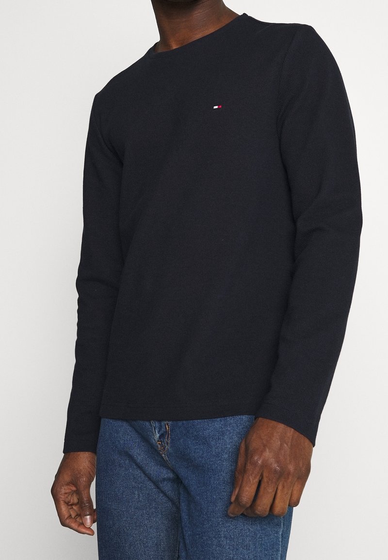 Schwarzes Langarm-T-Shirt aus strukturiertem Stoff, Rundhalsausschnitt und kleinem rot-weiß-blauem Logo auf der Brust. Kombiniert mit blauen Jeans.