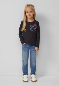 Młoda dziewczyna z długimi blond włosami, ubrana w czarną bluzkę z długim rękawem z uśmiechniętymi buźkami, niebieskie dżinsy i białe trampki, stojąca na jednolitym tle.