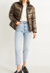 Person iført en brun pufferjakke, hvid skjorte, lyseblå højtaljede jeans og sorte loafers, stående mod en ensfarvet baggrund.