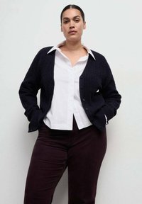 Femeie cu păr negru prins la spate, purtând o cămașă albă cu guler, un cardigan negru cu nasturi și pantaloni închiși la culoare, pe un fundal simplu, deschis la culoare.