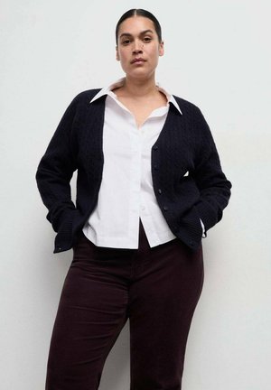 Femme aux cheveux foncés attachés, portant une chemise blanche à col, un cardigan noir boutonné et un pantalon sombre, sur un fond clair uni.