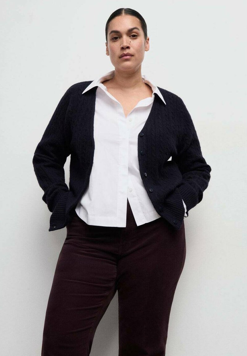 Femeie cu păr negru prins la spate, purtând o cămașă albă cu guler, un cardigan negru cu nasturi și pantaloni închiși la culoare, pe un fundal simplu, deschis la culoare.