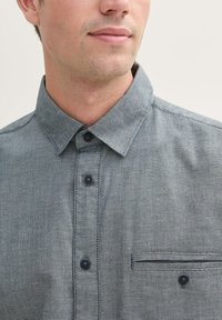 Graues, gemustertes Hemd mit Kragen, ausgestattet mit schwarzen Knöpfen und einer Brusttasche mit einem dekorativen Akzent am Taschenrand.