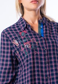 Camicia a scacchi blu navy con righe rosse e bianche, caratterizzata da ricami floreali dai colori vivaci e da una patta blu in contrasto. Bottoni visibili.
