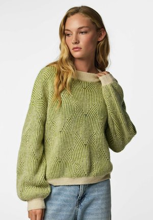 Femme aux longs cheveux blonds porte un pull ample vert à motifs avec des poignets crème et un jean bleu clair sur fond blanc uni.