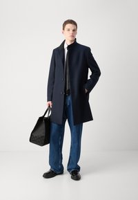 Cappotto navy con colletto alto, chiusura a un solo petto e tasche laterali, abbinato a jeans lunghi in denim e una borsa nera strutturata.