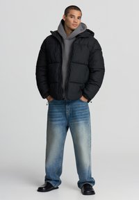 Czarna kurtka puchowa z kapturem, zapinana na zamek, o pikowanej strukturze, noszona na szarym hoodie i zestawiona z jasnoniebieskimi spodniami typu wide-leg.