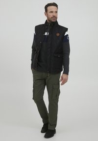 Fekete steppelt mellény szürke pulcsi felett, tengerészkék részletekkel, olívazöld cargo nadrággal és fekete cipővel párosítva; látható cipzárakkal és zsebekkel rendelkezik.