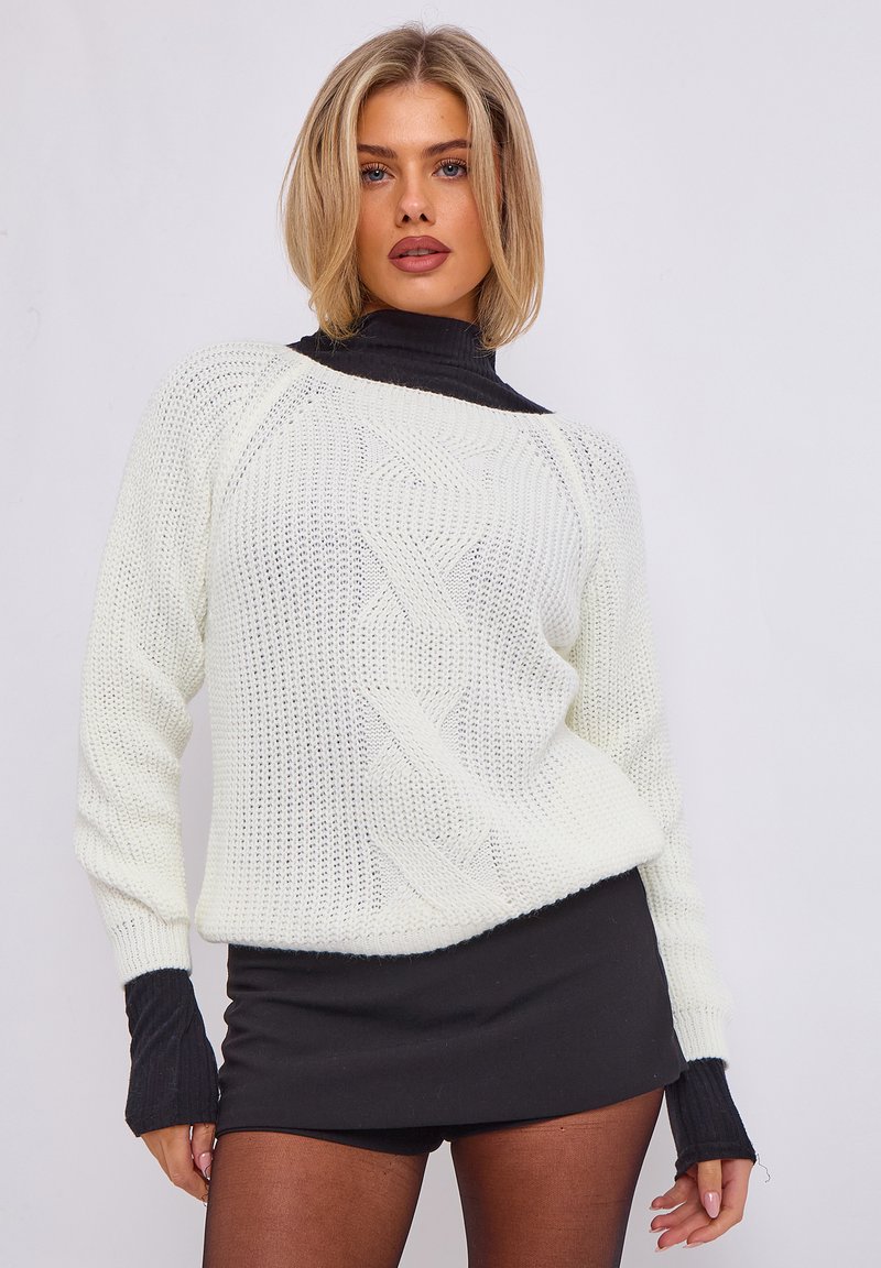 Weißer Strickpullover mit Zopfmuster und schwarzen Rippbündchen. Kombiniert mit einem schwarzen Rock, um einen Layering-Look zu kreieren.