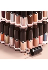 Filas de botellas de maquillaje líquido en varios tonos nude, marrón y metálico con tapas negras, una tapa quitada mostrando el aplicador.