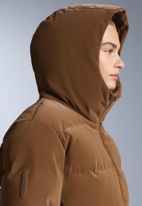 Smeđi pufasti kaput s kapuljačom, izrađen od glatkog, mat materijala. Ima zatvarač i RECCO® oznaku na lijevom rukavu.