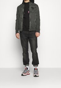 Man i blekt svart jacka, svart hoodie, mörka jeans som är upprullade vid fållen, vita strumpor och svarta, vita, rosa Nike-sneakers står mot en vit bakgrund.