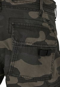 Pantalones cargo de camuflaje en verde oliva y negro. Presentan múltiples bolsillos y una textura en la tela. Incluye un pequeño lazo negro de sujeción.