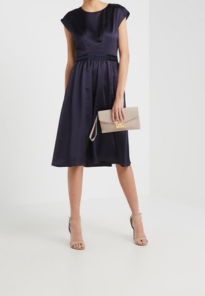 Mujer con vestido midi sin mangas de satén azul marino, sosteniendo un clutch beige y llevando sandalias de tacón alto color nude sobre un fondo blanco.