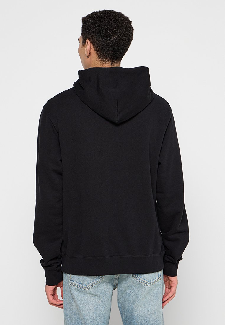 Schwarzer Kapuzenpullover mit langen Ärmeln. Hergestellt aus weichem Stoff, mit Kängurutasche und geripptem Saum. Getragen mit hellblauen Jeans.