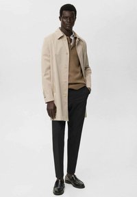 Man wearing beige trench coat over tan sweater, black pants, and black loafers, standing with one hand in pocket on plain background.