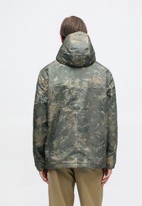 Maskáčová bunda v tlmených zelených a béžových odtieňoch, s kapucňou, elastickými manžetami a textúrovaným, vodeodolným vonkajším materiálom.