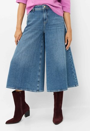 Person iført viddebenede denim culottes, en pink top og mørk burgunderrøde hælede ankelstøvler står mod en hvid baggrund.