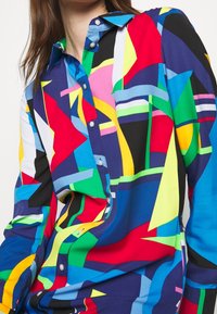 Flerfärgskjorta med knäppning framtill som har djärva geometriska mönster i röd, blå, gul, grön och svart. Slät tyg med klassisk krage.