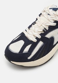 Marineblå og hvite sneakers med mesh- og semsket skinnmaterialer, et teksturert design og kremfargede snørebånd. Gummiyttersåle og synlige detaljer i aksent.