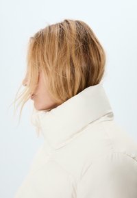 Puffjacka i krämfärg med hög krage. Slät textur och sydda paneler synliga på tyget. Håret är lätt rufsigt.