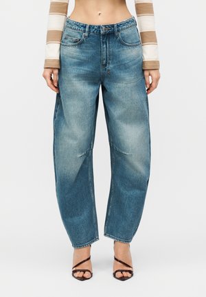 Γυναίκα φοράει φαρδιά, ξεβαμμένα μπλε τζιν με πέντε τσέπες, κουμπί και φερμουάρ, συνδυασμένα με καφέ σανδάλια με τακούνι και λουράκια.