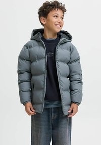 Grå puffjacka med huva, med quiltat mönster, dragkedjestängning och sidofickor. Bärs över en mörk sweatshirt och jeans.