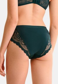 Culotte taille haute en teal avec une bordure en dentelle transparente et des bords festonnés, présentant des motifs floraux sur les côtés. Tissu lisse avec une finition mate.