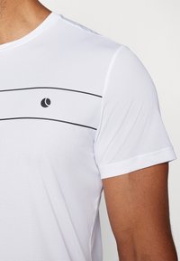 Valkoinen lyhythihainen urheilupaita, jossa on texturoitua kangasta, mustat vaakaraitat ja pieni logo rinnassa.