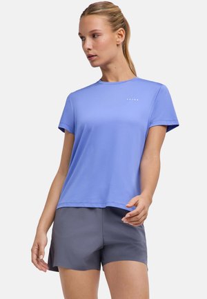 Frau mit blondem Haar im Pferdeschwanz, die ein hellblaues Kurzarmshirt und graue Shorts trägt, schaut nach links vor einem schlichten Hintergrund.