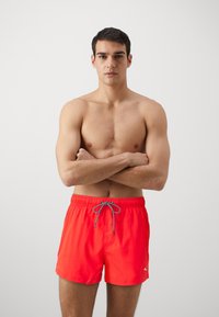 Red swim shorts with an elastic waistband and grey drawstring. Material appears lightweight; shorts feature a small logo on the side.