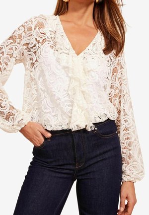 Woman wearing a white lace long-sleeve blouse with ruffle detail, paired with dark blue jeans, hand in pocket.