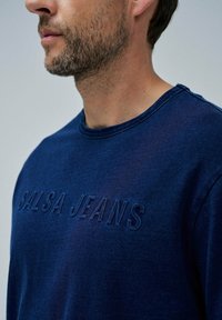 Námořnický modrý svetr vyrobený z měkké tkaniny, s vyšitým logem "SALSA JEANS", kulatým výstřihem a texturovaným povrchem.