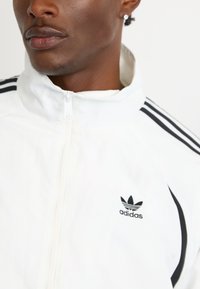 Zatvoren kadar osobe koja nosi bijelu Adidas trenirku s crnim prugama na ramenima i Adidas logotipom na prsima.