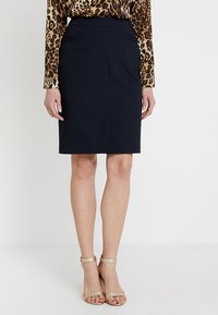 Jupe crayon bleu marine avec deux poches avant, tissu lisse, longueur genoux, associée à des talons dorés à lanières, portée avec un haut à imprimé léopard.