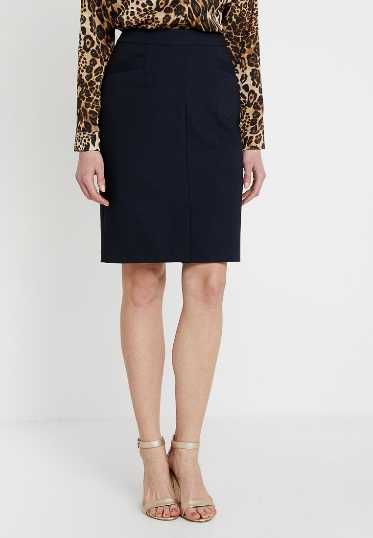 Jupe crayon bleu marine avec deux poches avant, tissu lisse, longueur genoux, associée à des talons dorés à lanières, portée avec un haut à imprimé léopard.