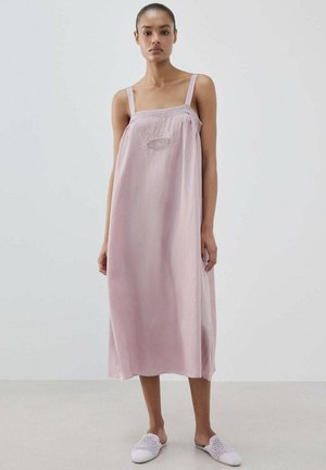 Mujer con un vestido midi sin mangas de color rosa claro con bordados en el pecho y zapatos blancos tejidos tipo slip-on, posando frente a un fondo sencillo.