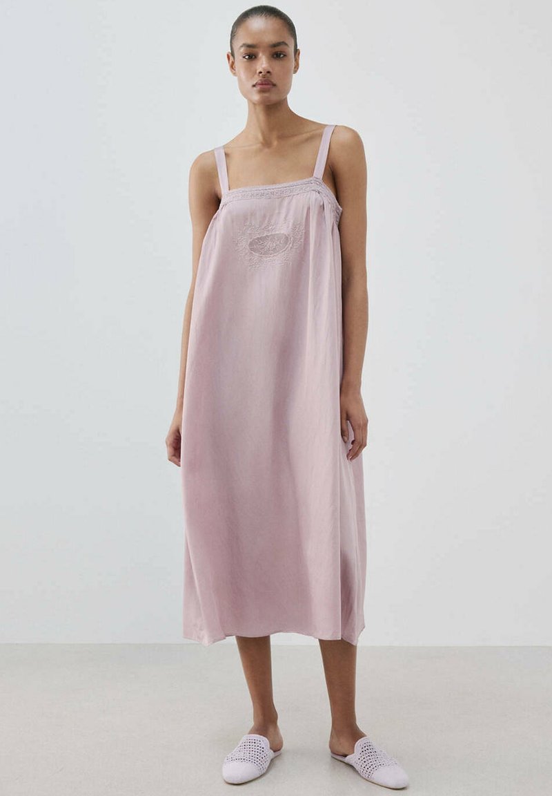 Femme portant une robe midi sans manches rose clair avec une broderie sur la poitrine et des chaussures blanches tissées à enfiler, debout devant un fond uni.