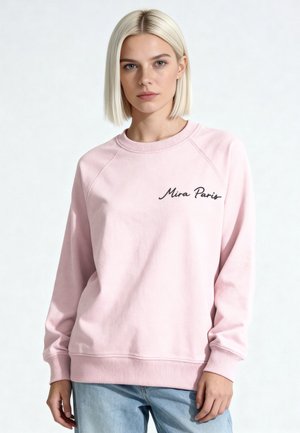Jonge vrouw met blond bobkapsel die een lichtroze trui met ronde hals draagt met de tekst "Mira Paris" en lichtblauwe spijkerbroek, staand voor een witte achtergrond.