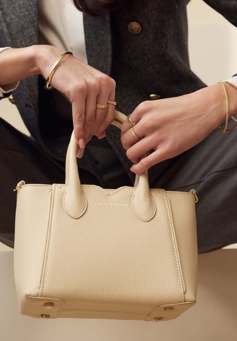 Beige Lederhandtasche mit kurzen Griffen, strukturiertem Material und goldenen Beschlägen. Markenname auf der Vorderseite eingeprägt; minimalistisches Design.