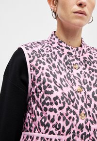 Ροζ καπιτονέ γιλέκο με μαύρο λεοπάρ σχέδιο, με όρθιο γιακά, εμφανείς κουμπιά και ένα αντίθετο μαύρο μακρυμάνικο μπλουζάκι από κάτω.