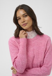 Suéter de punto rosa de textura suave con mangas largas. Llevado sobre una blusa blanca con volantes, con un escote redondo y detalles de botones.