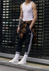 Muž ve bílém tílku, černých sportovních kalhotách Adidas, bílých teniskách a hnědé kockované košili uvázané kolem pasu, stojící venku.