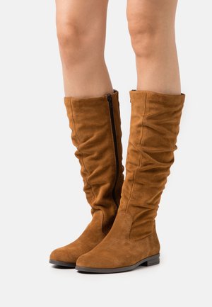 Bottes en daim marron à hauteur de genou avec un design plissé et fermeture éclair noire sur le côté. Semelle plate et bout rond avec une texture lisse.
