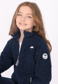 Giacca a zip in pile blu navy con colletto alto, logo bianco a forma di ombrello sul petto e una toppa rotonda sulla manica. Tessuto texturizzato.