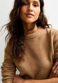 Light brown wool sweater with a ribbed crew neckline and long sleeves, worn by a person with wavy hair against a neutral background.