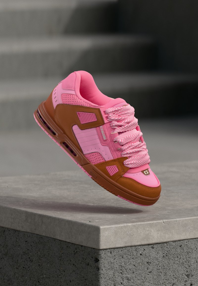 Pantof sport pink și maro cu șireturi groase roz plutind deasupra unei margini de beton, având în fundal trepte gri neclare.