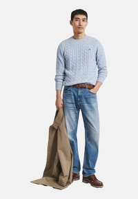 Helles blaues Cable-Knit-Pullover, blaue Denim-Jeans, braune Lederstiefel, braner Gürtel, hält einen hellbraunen Mantel. Einfaches und lässiges Outfit.