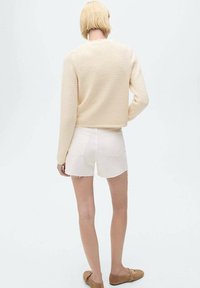 Suéter de punto recortado beige con tejido texturizado, combinado con pantalones cortos de mezclilla blanca con dobladillos deshilachados y bolsillos traseros. Zapatos marrones completan el look.